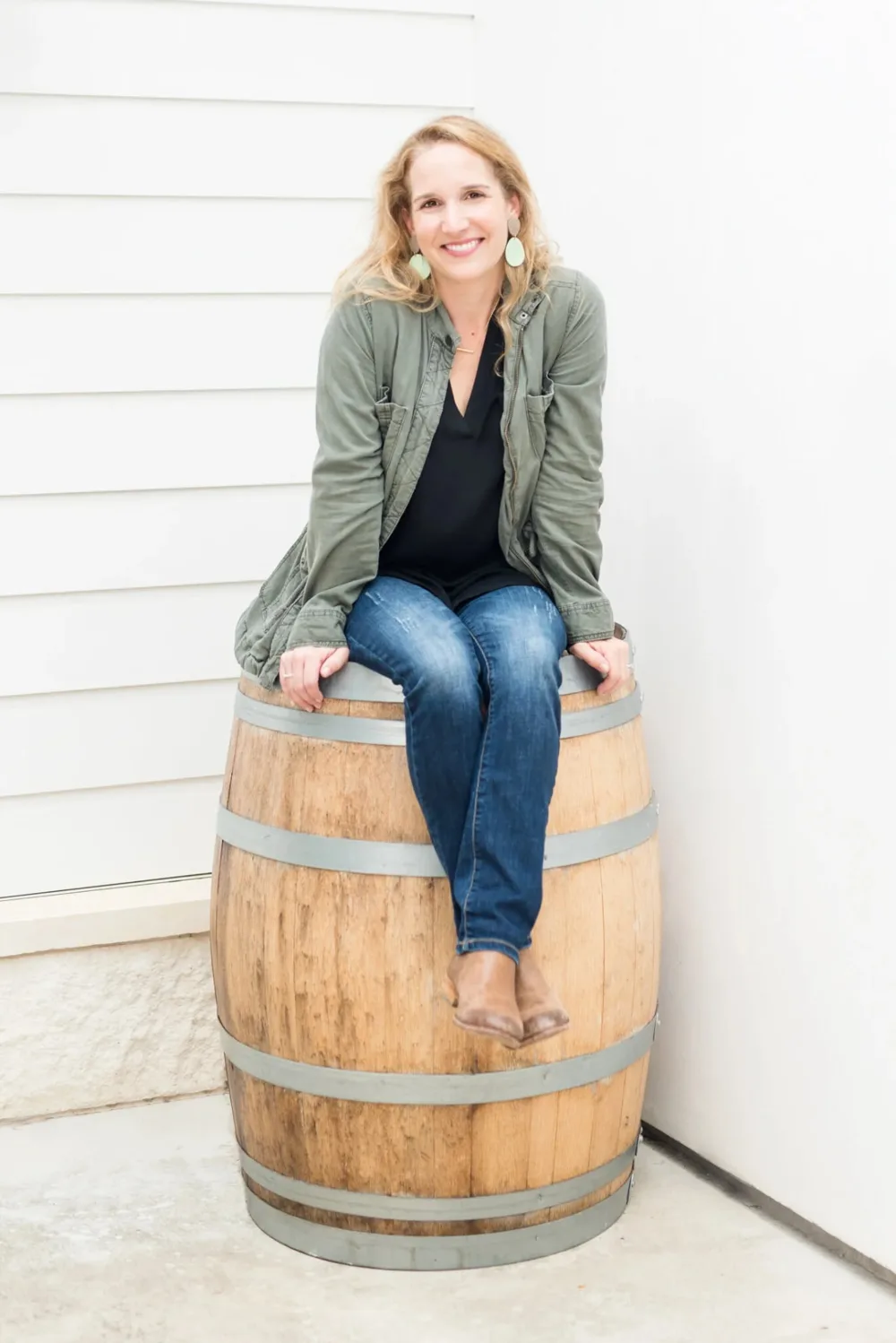 Jessica Dupuy smiling, seated on an oak wine barrel in a green jacket and jeans.